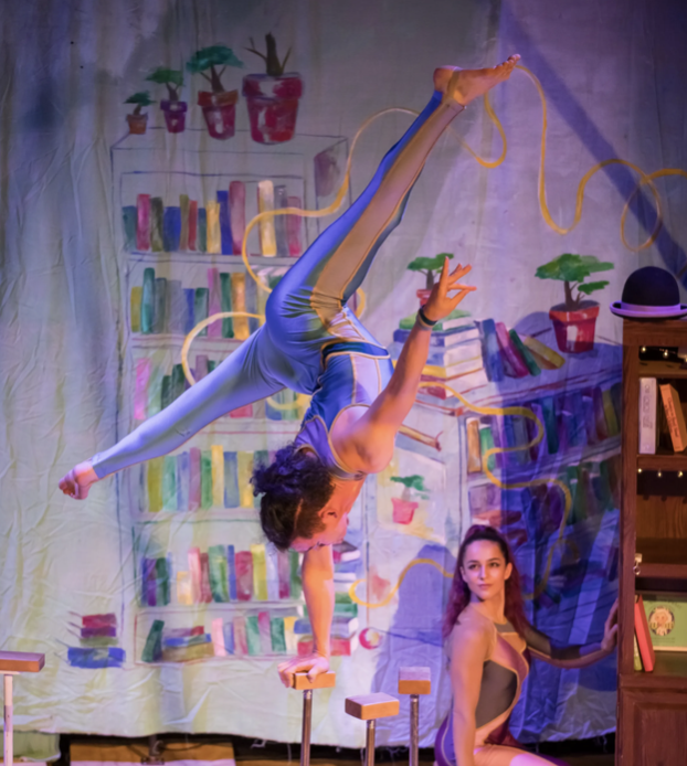 A male-presenting acrobat performs a handstand on cylindrical props against a colorful library backdrop, while a female-presenting performer watches.