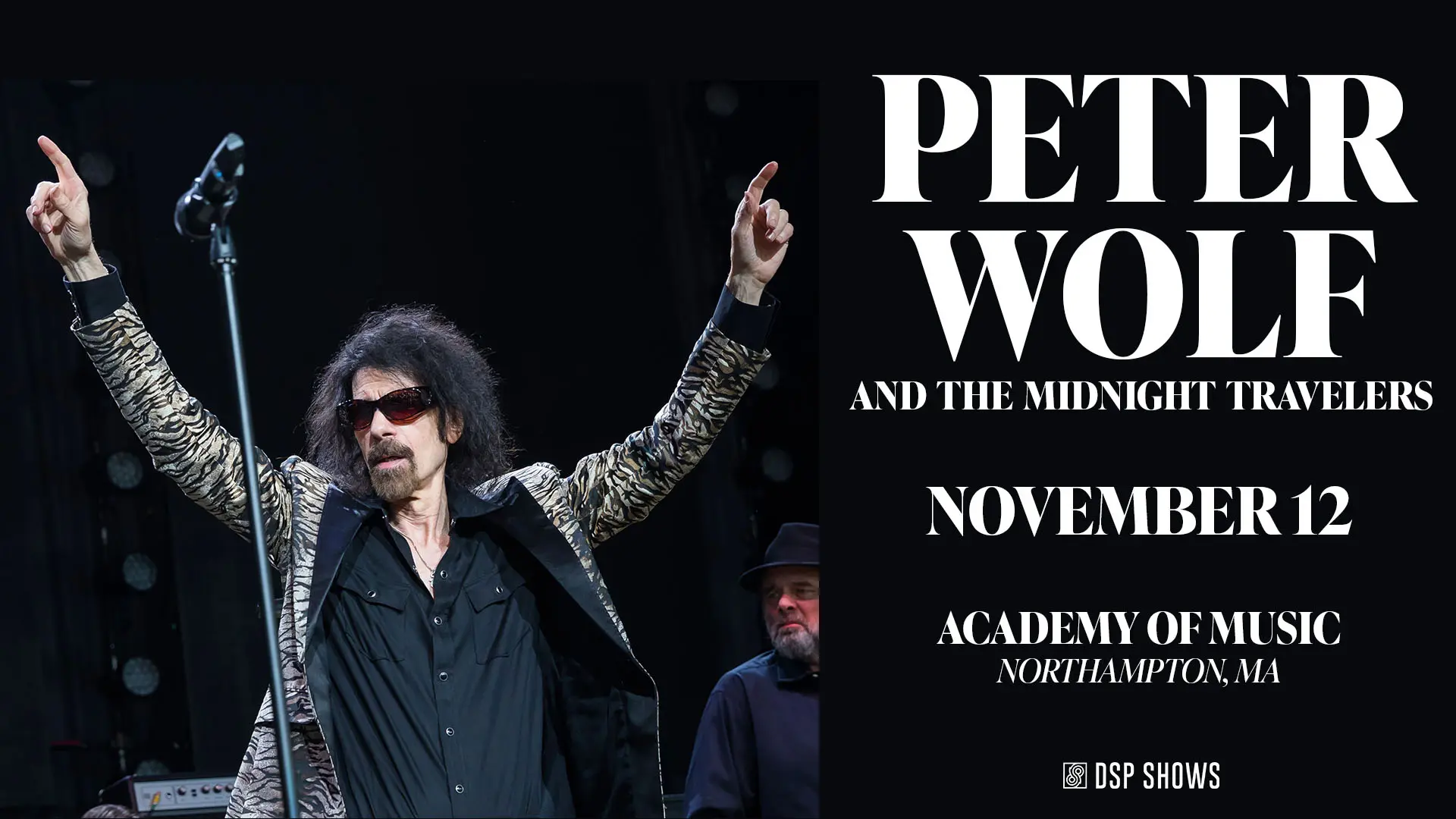 Peter Wolf performing on stage, wearing sunglasses and a patterned jacket, with arms raised while engaging with the audience.