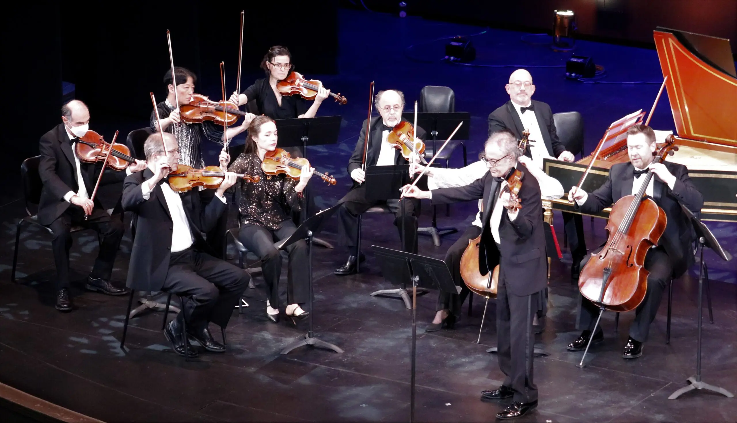 A classical music ensemble of nine musicians in formal black attire performs on stage, seated in a semi-circle with violins and a cello, against a blue backdrop.
