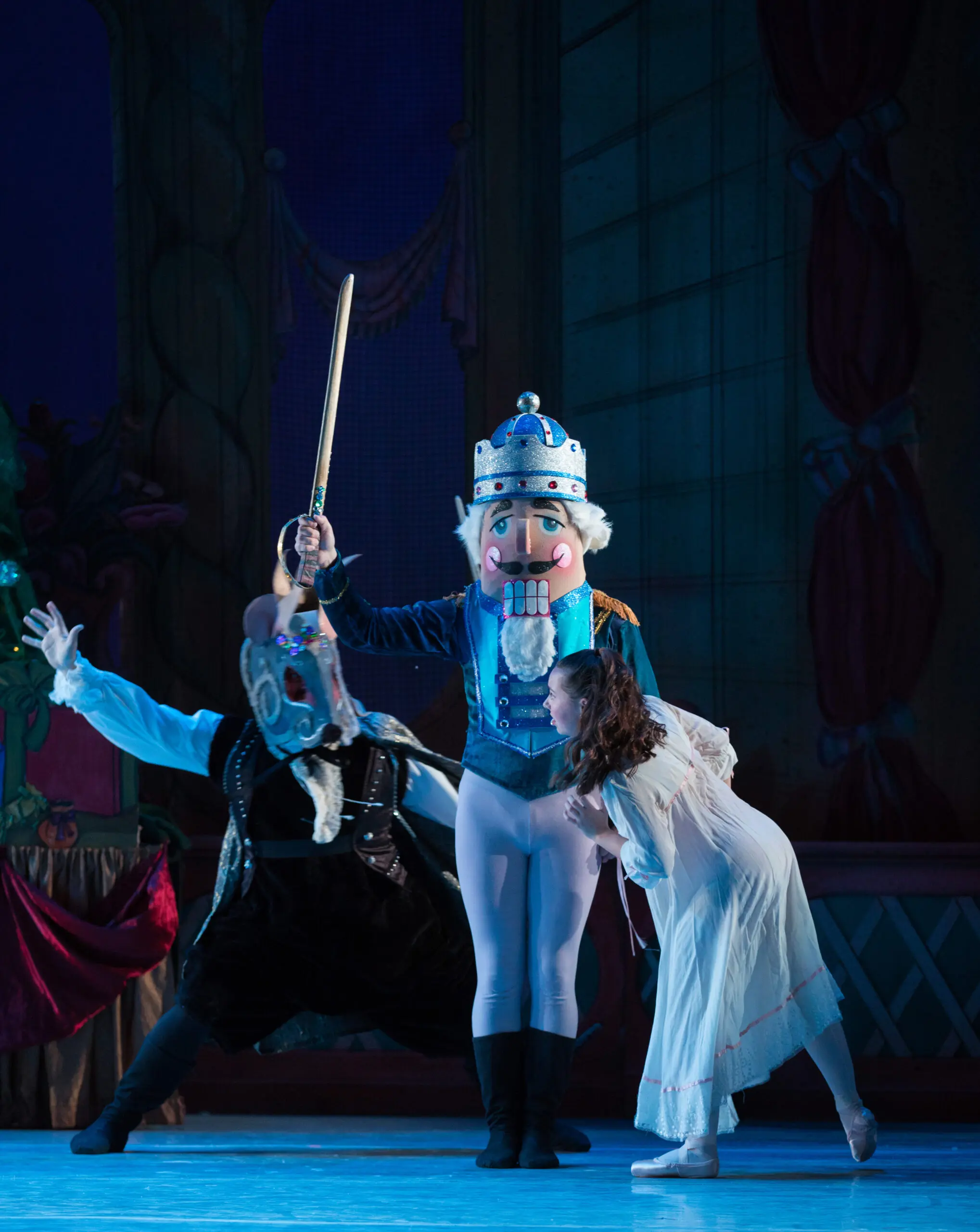 A person in a nutcracker costume stands in the middle with a sword raised up, defending a young girl in a nightgown from a person in a rat costume