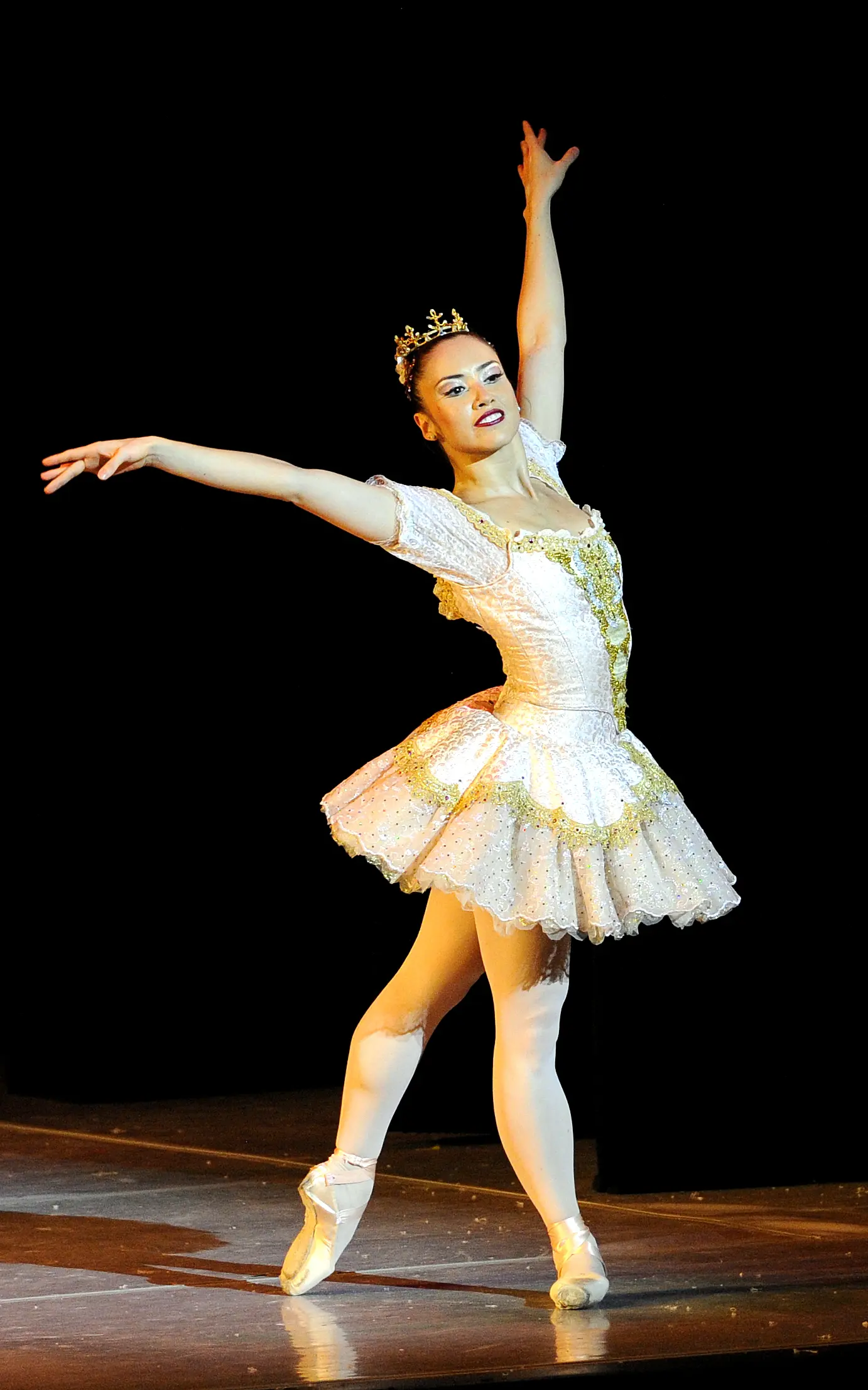 A Ballerina dressed in a white tutu on pointe shoes with their arms extended upwards.