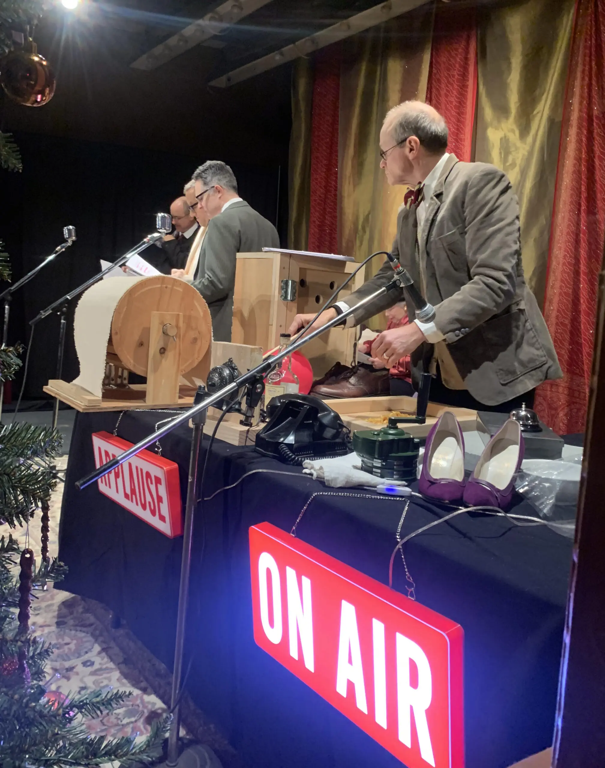 Three men perform on a decorated stage behind microphones, wooden instruments, and a table of props; red signs read “ON AIR” and “PLEASE.”
