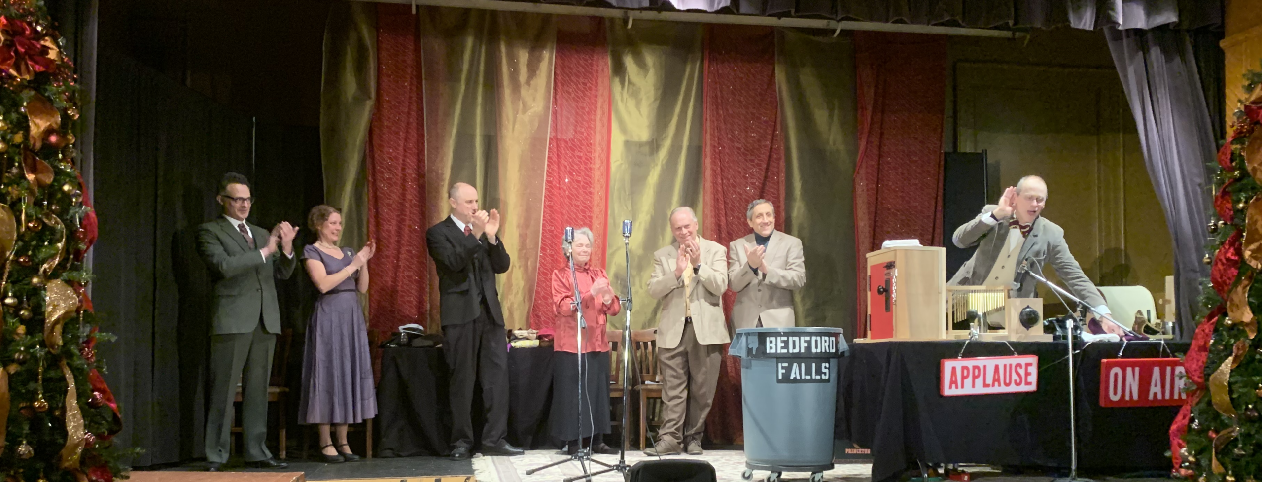 Seven performers applaud on a Christmas-decorated theater stage beside vintage microphones and broadcast props labeled “BEDFORD FALLS,” “APPLAUSE,” and “ON AIR.”