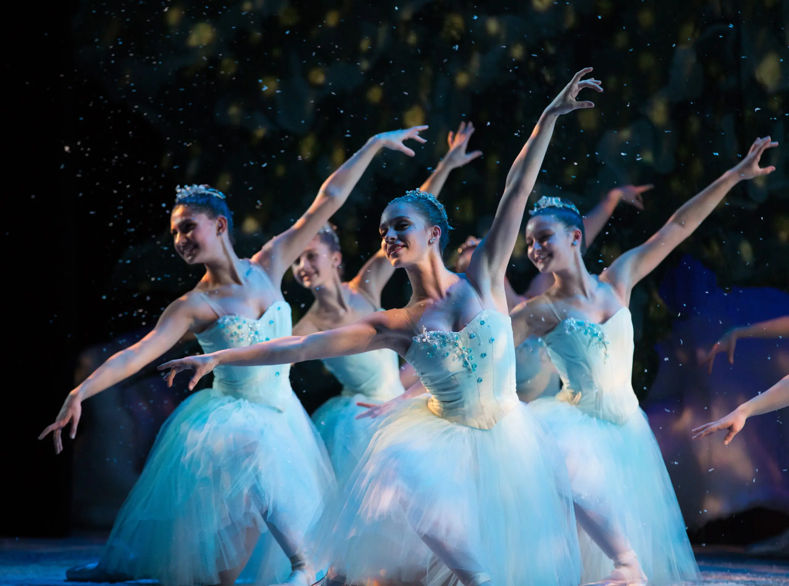 Four ballerinas dressed in white with their left arms extended upwards.