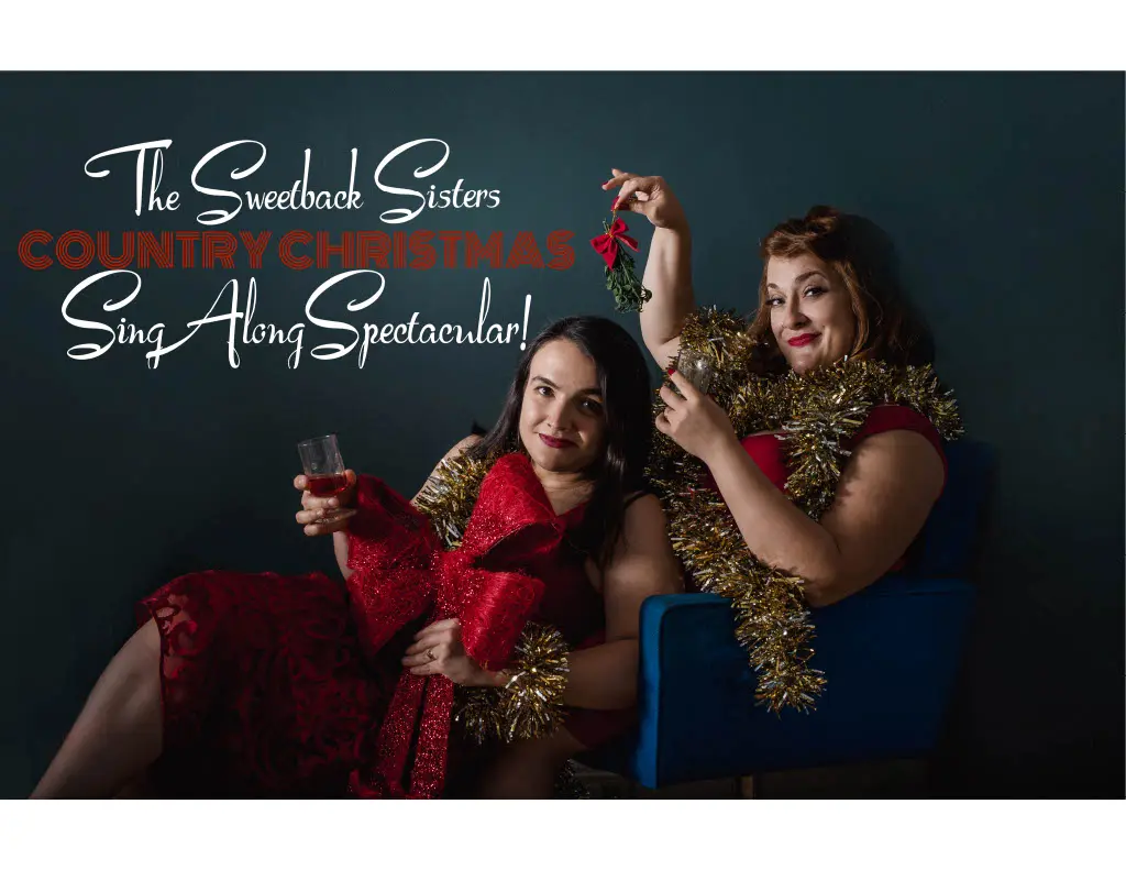 Two feminine presenting people. One sitting down with dark hair and holding a drink while the other is sipping a drink while holding a sprig of mistletoe above the duo.