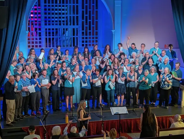 Large group choir performing on a brightly lit stage while a conductor and audience watch.